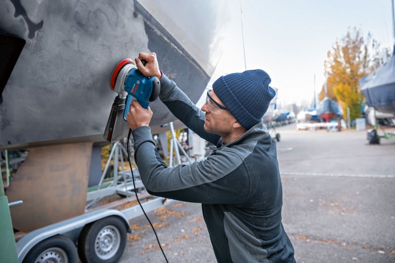 A person wearing safety equipment sands a boat hull outdoors with a random orbit sander.