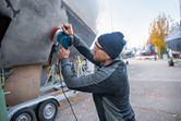 A person wearing safety equipment sands a boat hull outdoors with a random orbit sander.