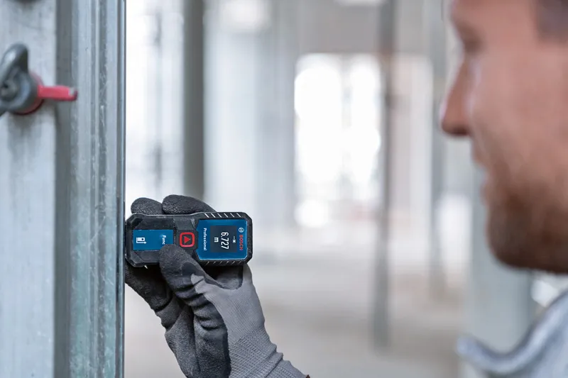 A person wearing safety equipment uses a laser measure to check distance on a metal frame.