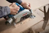A person cuts a wooden board with a cordless circular saw, creating sawdust.