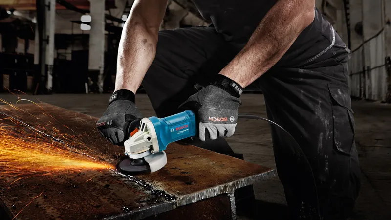 A person wearing safety equipment grinds a metal beam with an angle grinder, producing sparks.