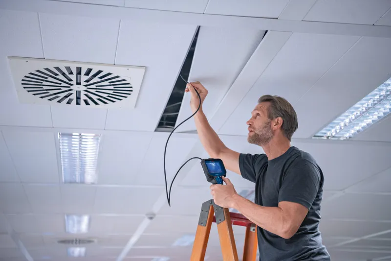 A person on a ladder inspects a ceiling cavity with an inspection camera.
