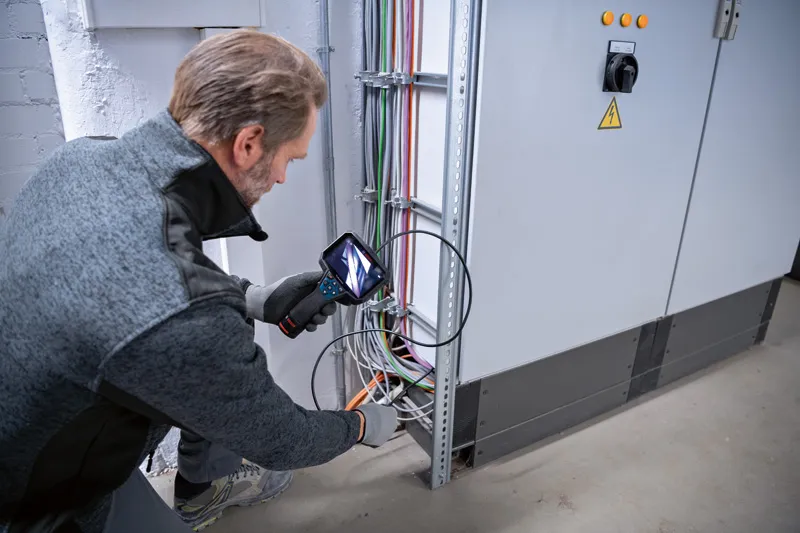 A person wearing safety equipment inspects electrical wiring using an inspection camera.