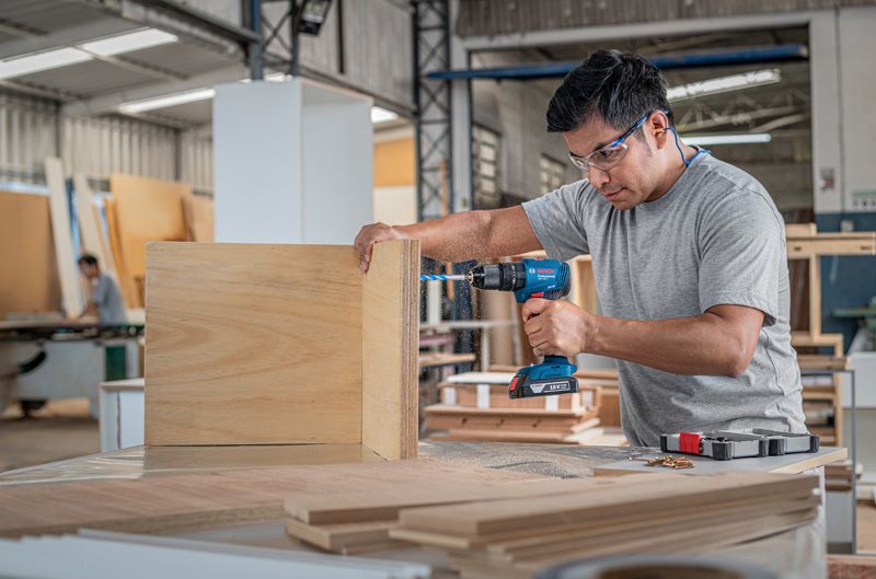 A person wearing safety equipment drills holes in wooden boards in a workshop.