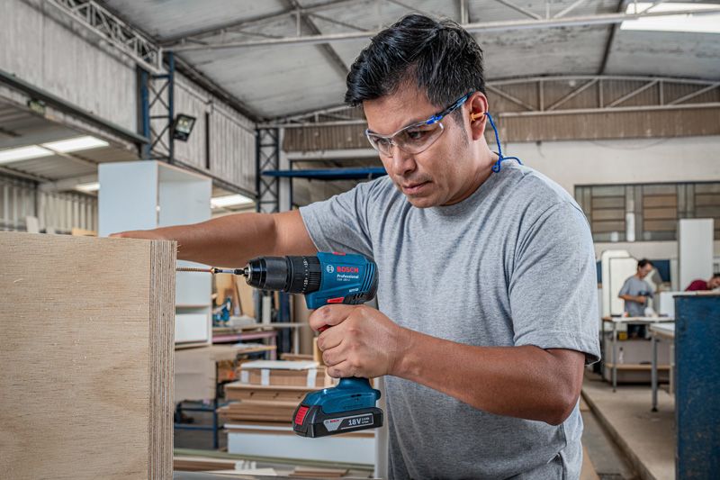 A person wearing safety equipment drills into plywood in a woodworking shop.
