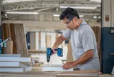 Person wearing safety equipment uses a cordless drill on wooden boards in a workshop.