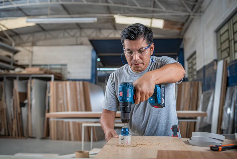 A person wearing safety equipment drills a hole into wood in a workshop.