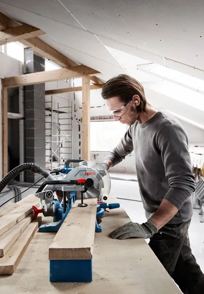 A person wearing safety equipment cuts a wooden board with a circular saw in a workshop.