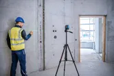 A person wearing safety equipment marks a concrete wall using a laser leveling tool on a tripod.