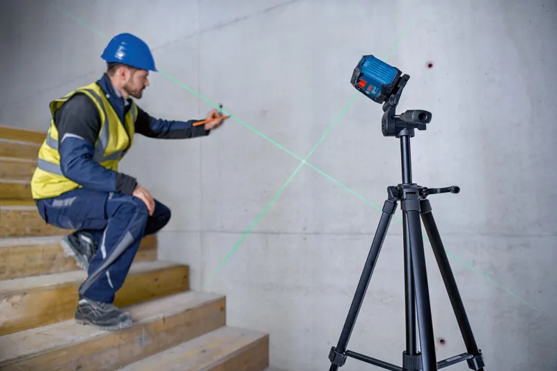 A person wearing safety equipment marks a wall using a green laser leveling tool on a tripod.