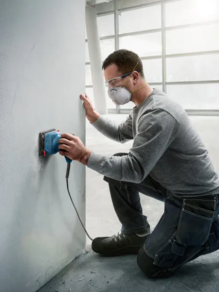 A person wearing safety equipment sands a wall with an orbital sander.