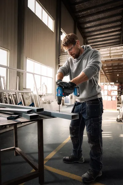 A person wearing safety equipment drills into a metal beam in a workshop.