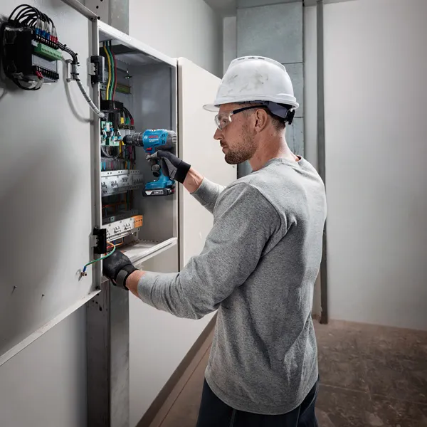A person wearing safety equipment uses a cordless drill on an electrical panel.