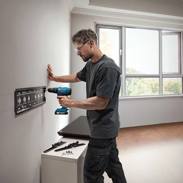 A person wearing safety equipment drills holes in a wall to mount a metal bracket.