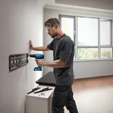 A person wearing safety equipment drills holes in a wall to mount a metal bracket.