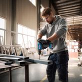 A person wearing safety equipment drills into a metal beam inside a workshop.