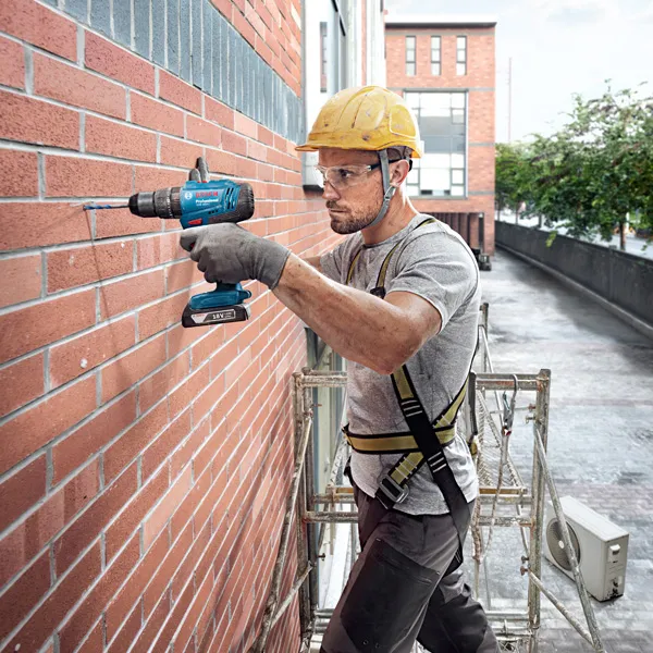 A person wearing safety equipment drills into a brick wall on scaffolding.