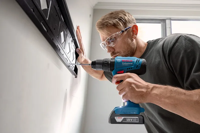 A person wearing safety equipment drills into a wall to install a mounting bracket.