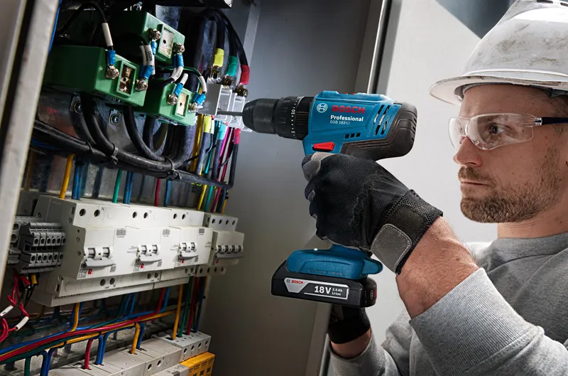 A person wearing safety equipment drills into a control panel full of wires.