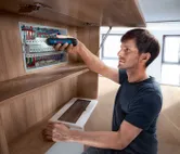 A person uses a cordless screwdriver to fasten components in an electrical panel.