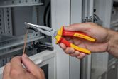 A person grips insulated pliers, shaping a wire near an electrical panel.