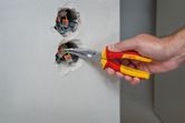 A person uses insulated bent nose pliers to adjust electrical wiring in a wall socket.