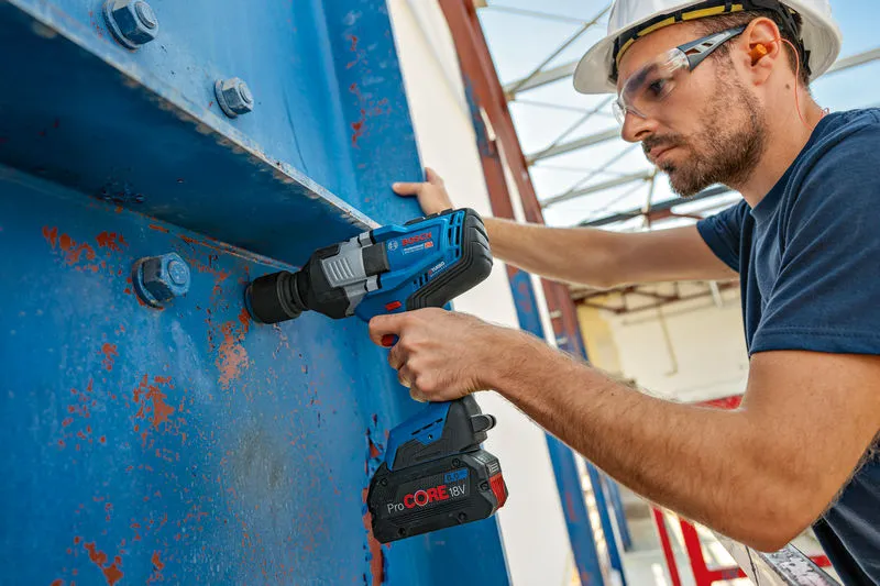 A person wearing safety equipment uses a cordless impact wrench on a large metal beam.