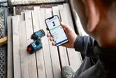 A person checks a power tool's app features on a smartphone near a cordless drill.