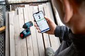 A person checks a power tool's app features on a smartphone near a cordless drill.