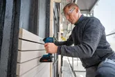 A person wearing safety equipment uses a cordless drill to fasten wooden boards outdoors.