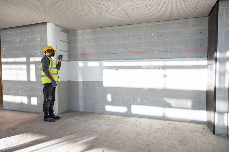 A person wearing safety equipment uses a laser leveling tool to project a straight line on a wall.