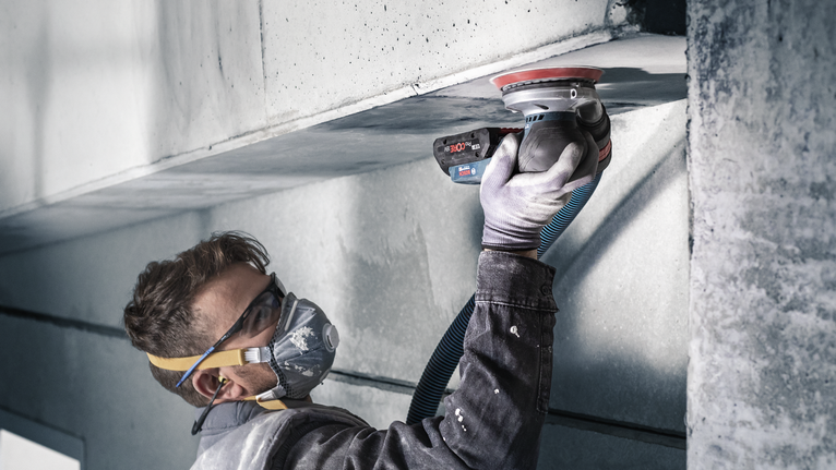 Person wearing safety equipment uses a power sander on a concrete ceiling.