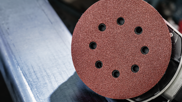 A disc sander with a red abrasive pad is positioned above a metal surface.