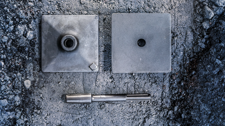 Metal core bit and two square mounting plates on a rough concrete surface.