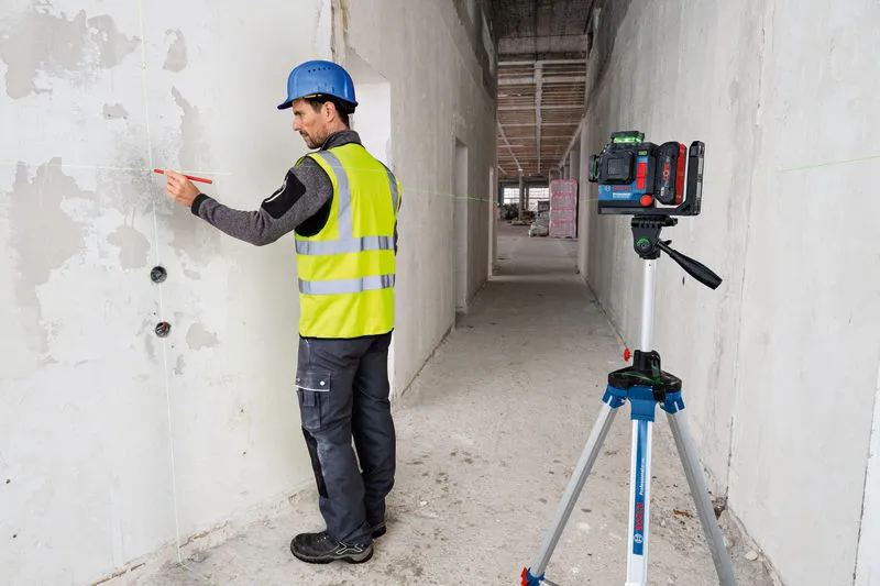 A person wearing safety equipment marks a wall guided by a laser leveling tool on a tripod.