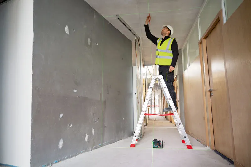 A person wearing safety equipment marks a ceiling using a laser leveling tool.