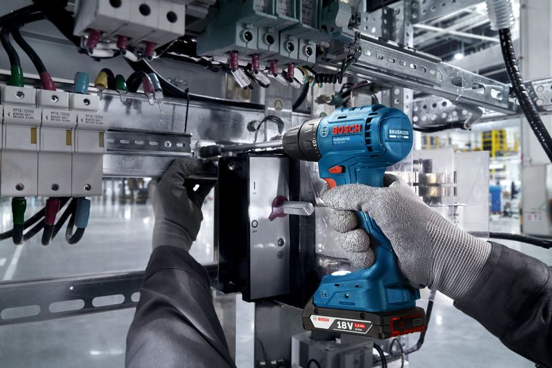 A person wearing safety equipment uses a cordless drill on an electrical panel.