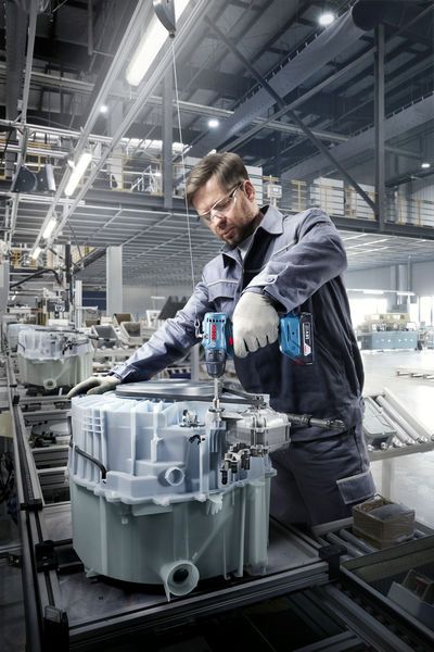 A person wearing safety equipment uses a cordless drill to assemble machinery in a factory.