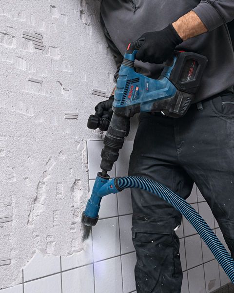A person wearing safety equipment uses a cordless demolition hammer to break wall tiles.