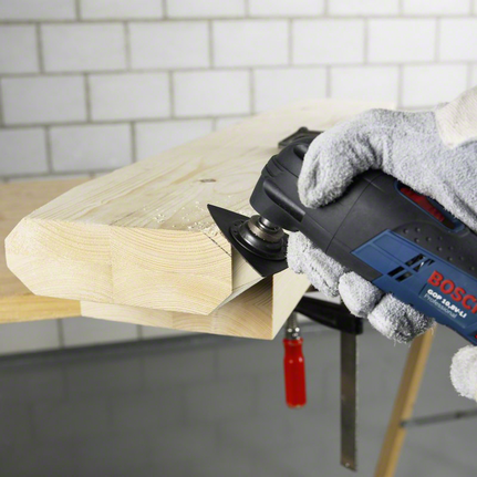 Gloved worker wearing safety equipment trims a wooden edge with a multi-tool.