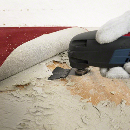 Person wearing safety equipment uses a multi-tool to remove adhesive under flooring.