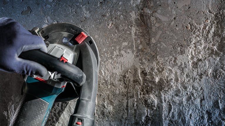 Person wearing safety equipment operates a power tool on a rough concrete wall.