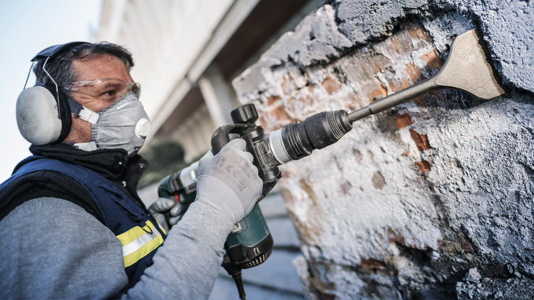 Person wearing safety equipment chisels a brick wall with a power tool.