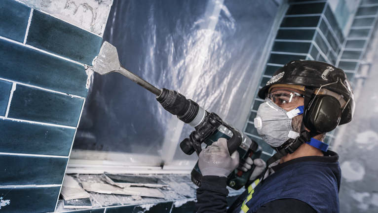Person wearing safety equipment uses a power tool to remove ceramic tiles from a wall.