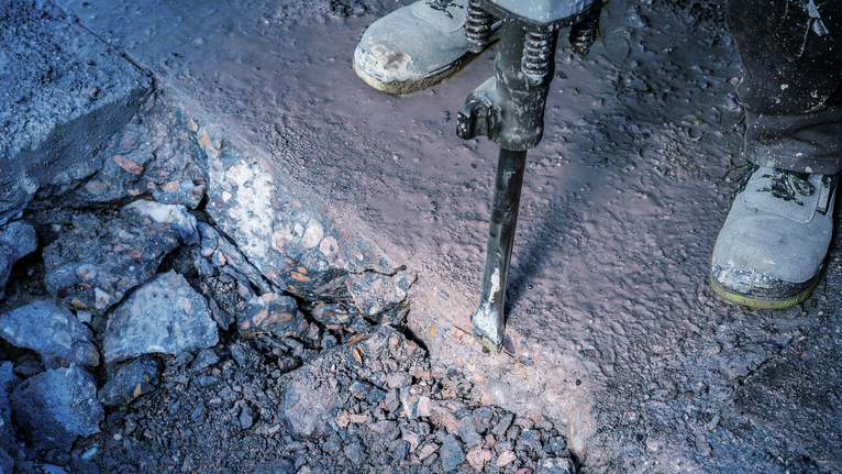 Person wearing safety equipment breaks concrete with a demolition tool.