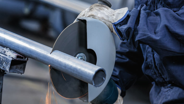 A person wearing safety equipment cuts a metal pipe with a circular saw.