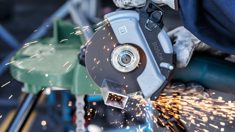 Person wearing safety equipment cuts metal pipe with a circular saw, creating sparks.