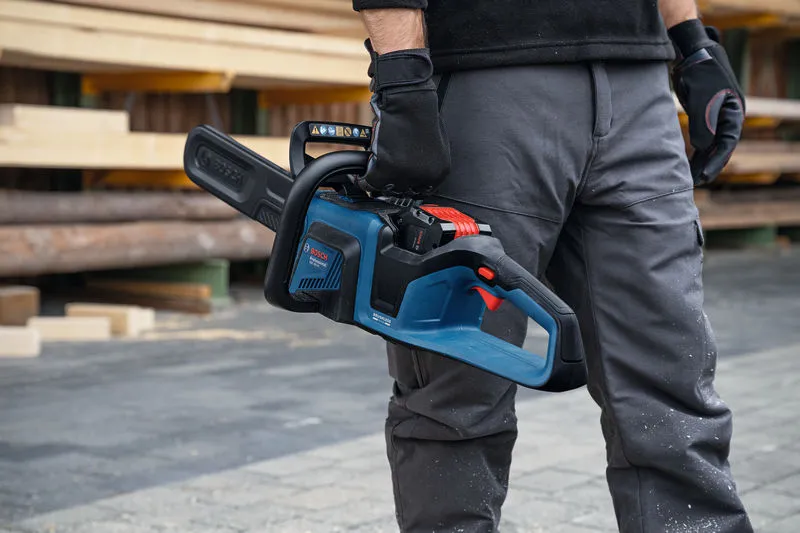 A person wearing safety equipment holds a cordless chainsaw at a lumber yard.
