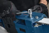 A person wearing safety equipment adjusts a cordless chainsaw with a wrench.