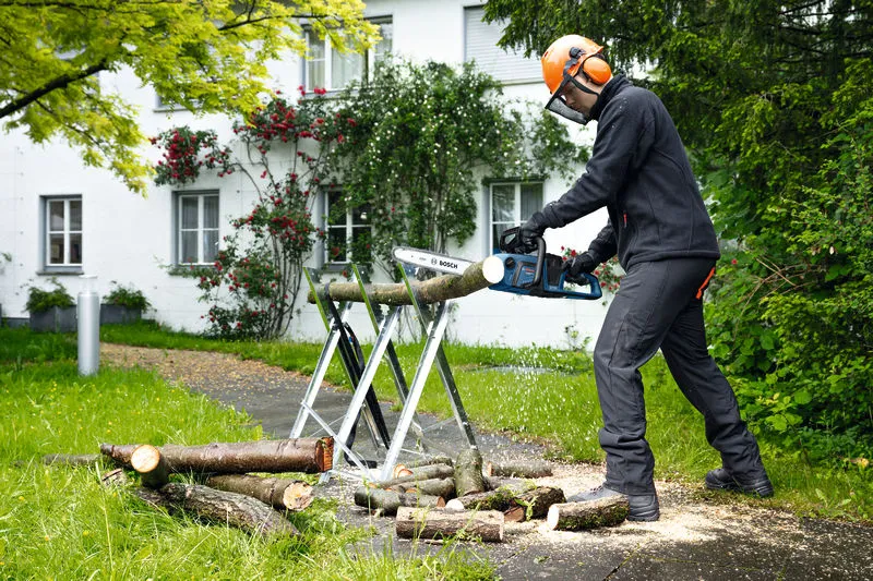 A person wearing safety equipment uses a cordless chainsaw to cut logs outdoors.